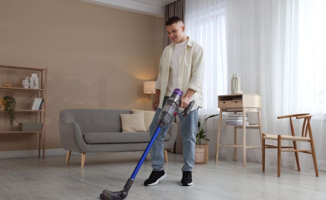 Un homme passant un aspirateur multifonction balai bleu sur un parquet clair dans un salon au design épuré.