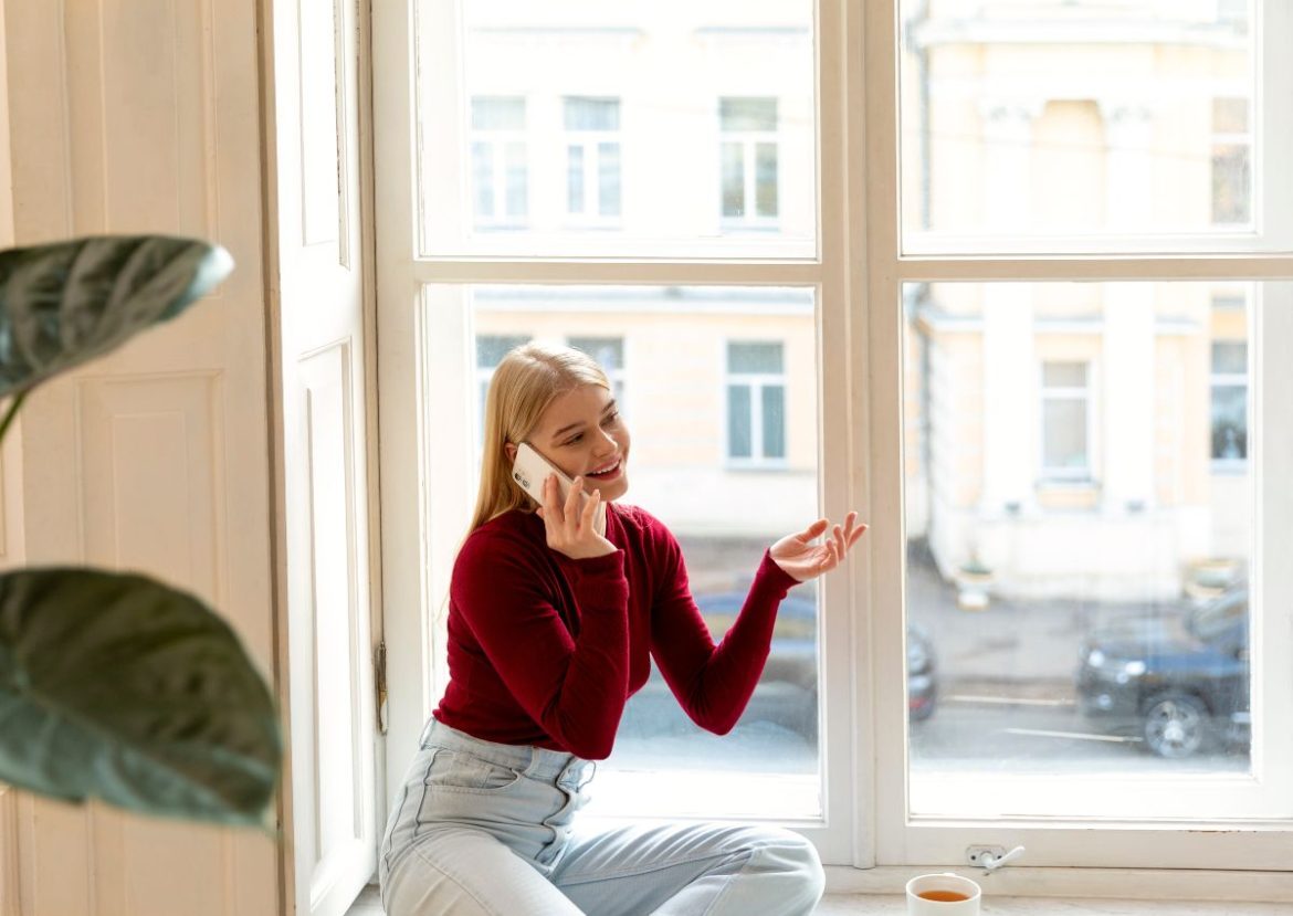 Jeune femme blonde souriante en pull rouge, assise sur le rebord d'une fenêtre à Montpellier et discutant au téléphone, avec vue sur un bâtiment ancien.