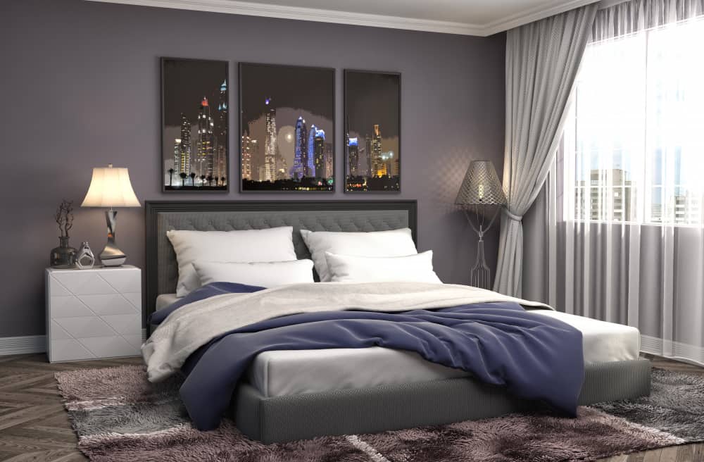 Décoration moderne d'une chambre parentale avec un grand lit double en tissu gris, des murs mauve foncé et un triptyque représentant une ville illuminée la nuit.