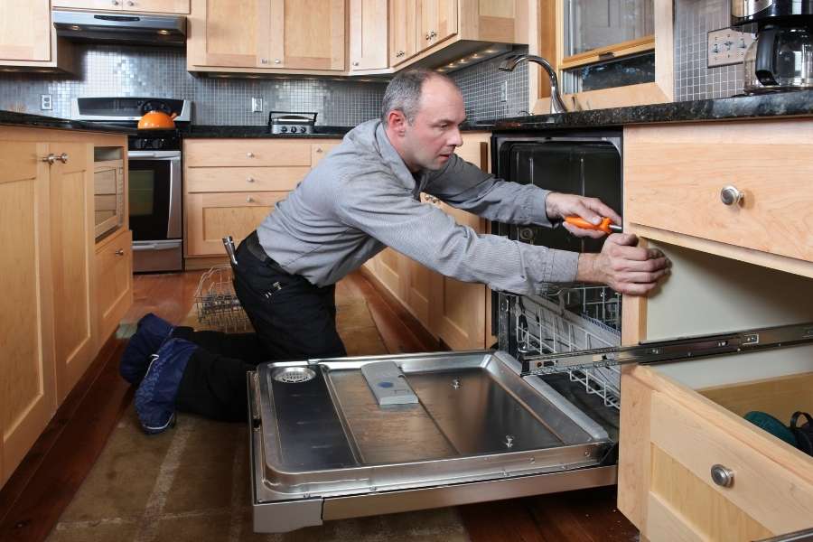 Un technicien professionnel, portant des sur-chaussures de protection, est agenouillé dans une cuisine équipée et utilise un tournevis pour ajuster le panneau d'habillage d'un lave-vaisselle encastrable.
